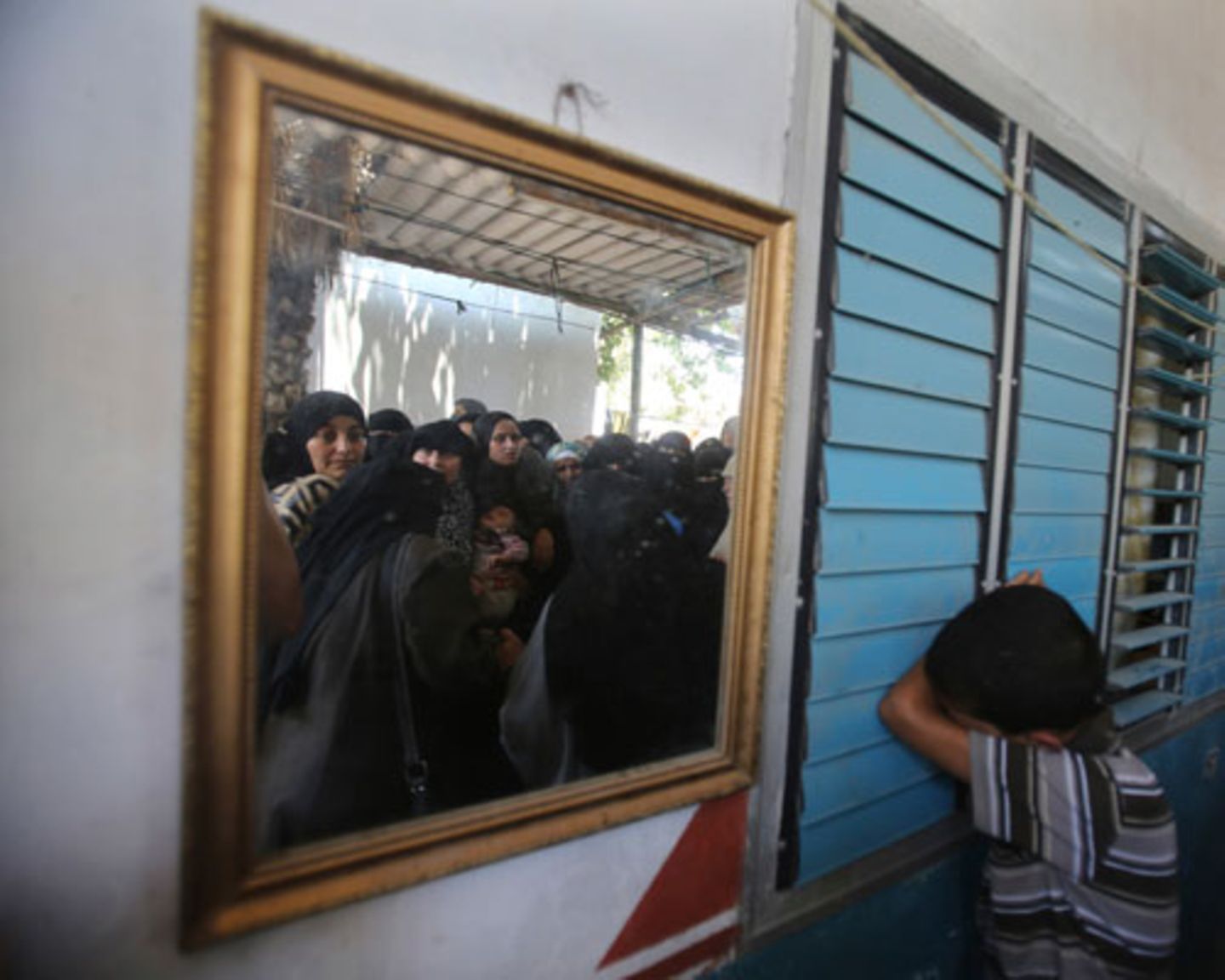 Ein palästinensischer Junge trauert um das vierjährige Mädchen Sarah Sheik al-Eid, das zusammen mit ihrem Vater und ihrem Onkel bei Luftangriffen getötet wurde. Im Spiegel sind Frauen zu sehen, die ebenfalls an der Trauerfeier in der Stadt Rafah teilnehmen.