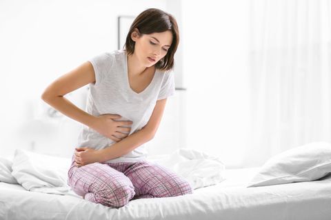 Medizin: Frau sitzt auf dem Bett und hält sich unter Schmerzen den Bauch