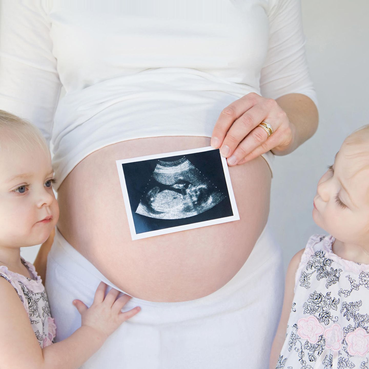 Geschwisterkinder stehen neben Babybauch und schauen darauf gehaltenes Ultraschallbild an.