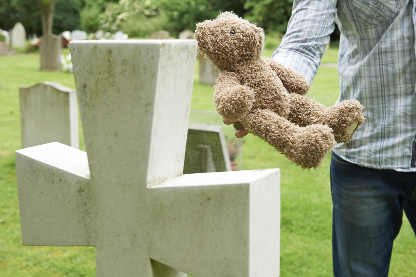 Vater steht mit einem Stoff-Teddy vor einem Grab