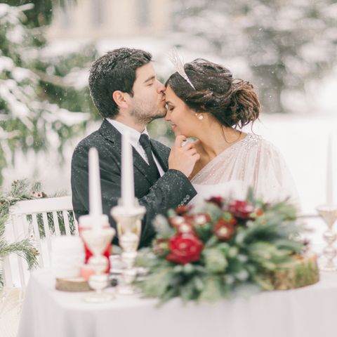Winterhochzeit - Tipps für deinen Traum in Weiß: Brautpaar sitzt draussen bei Schnee am geschmückten Tisch, er küsst sie auf