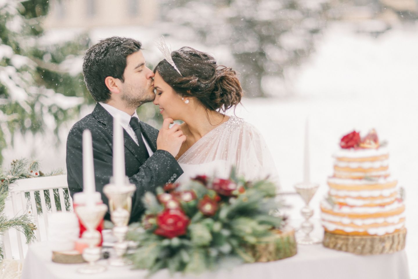 Winterhochzeit - Tipps für deinen Traum in Weiß: Brautpaar sitzt draussen bei Schnee am geschmückten Tisch, er küsst sie auf