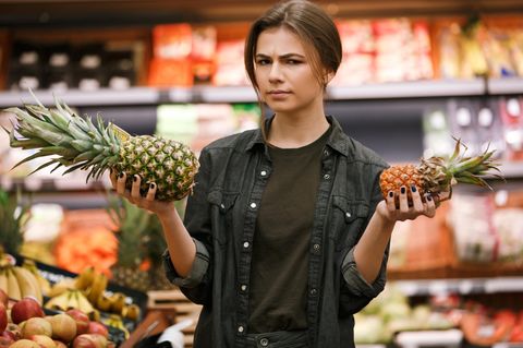 Junge Frau hält im Supermarkt skeptisch eine Ananas hoch