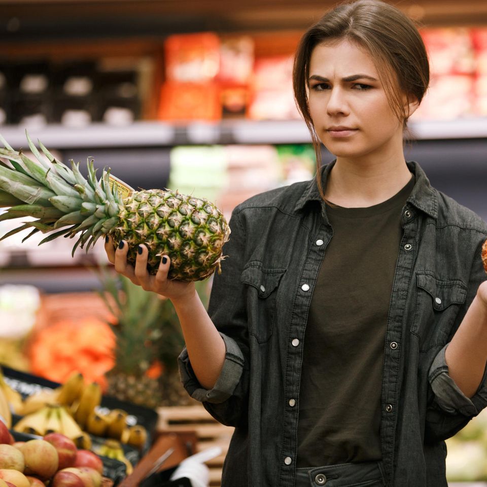 Junge Frau hält im Supermarkt skeptisch eine Ananas hoch