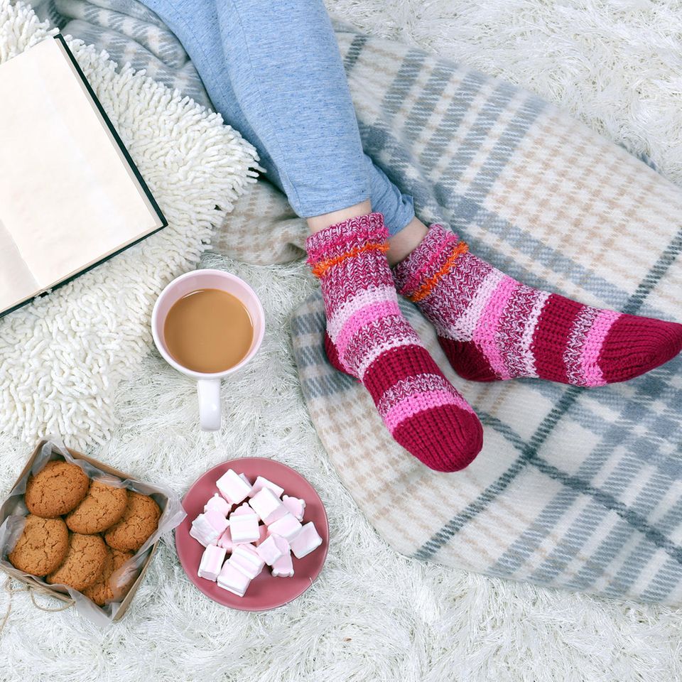 Tipps bei Kälte: Frau sitzt mit selbstgestrickten Socken an den Füßen auf dem Bett