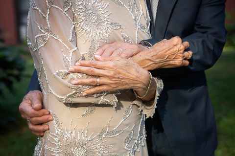 Steinerne Hochzeit: Bedeutung, Sprüche und Geschenkideen: Älteres Ehepaar in Festagsrobe umarmen sich und zeigen Eheringe i