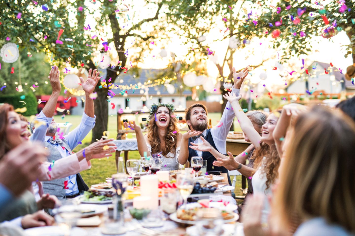 Gartenhochzeit - Tipps und Ideen für eure Traumhochzeit: Brautpaar sitzt mit Gästen im Garten am Tisch und werfen Konfetti