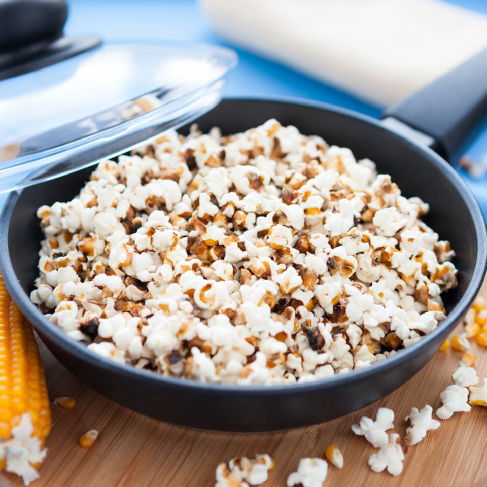 Popcorn selber machen: So leicht geht das Rezept: Pfanne gefüllt mit Popcorn steht auf einem Brett, ein Maiskolben liegt daneben