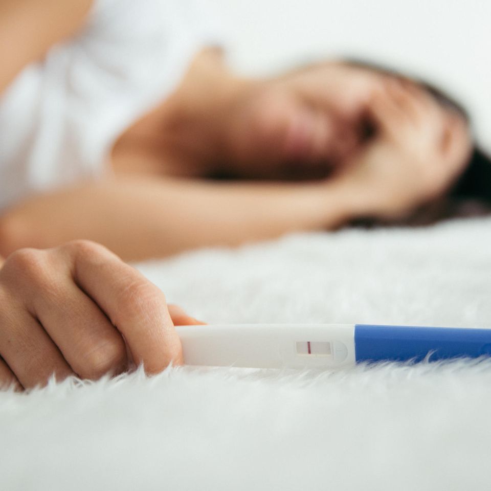 Unerfüllter Kinderwunsch: Frau liegt mit negativem Schwangerschaftstest auf weißem Teppich