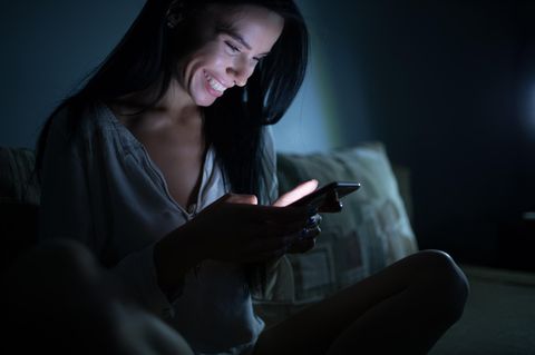 Romance Scamming: Eine junge Frau im dunklen Schlafzimmer mit Handy