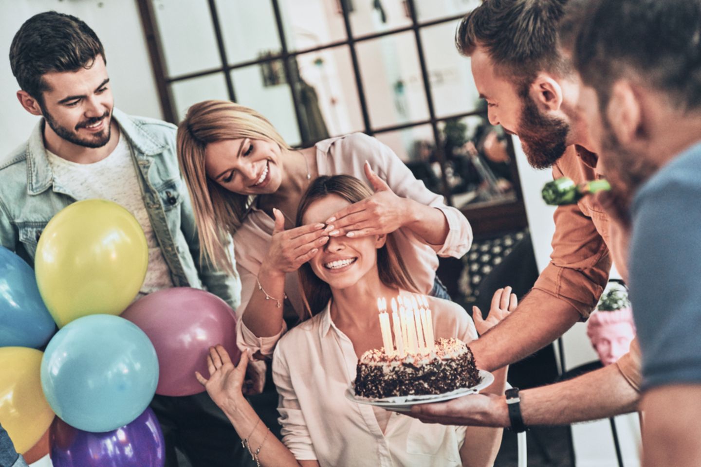 Überraschungsparty: Tipps, wie der Plan aufgeht! Frau werden die Augen zugehalten, vor ihr eine Geburtstagstorte mit Kerzen