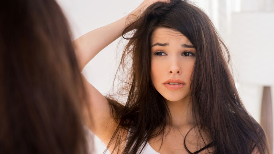 Verfilzte Haare: 6 effektive Tipps für kämmbares Haar: Frau fasst sich in die langen Haare und schaut unglücklich in den Spiegel