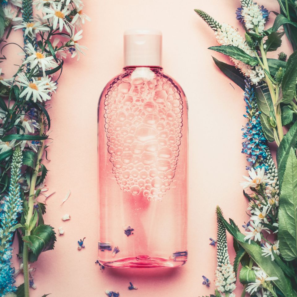Gesichtswasser selber machen: Flasche mit durchsichtiger Flüssigkeit auf rosa Untergrund, umgeben von Blumen