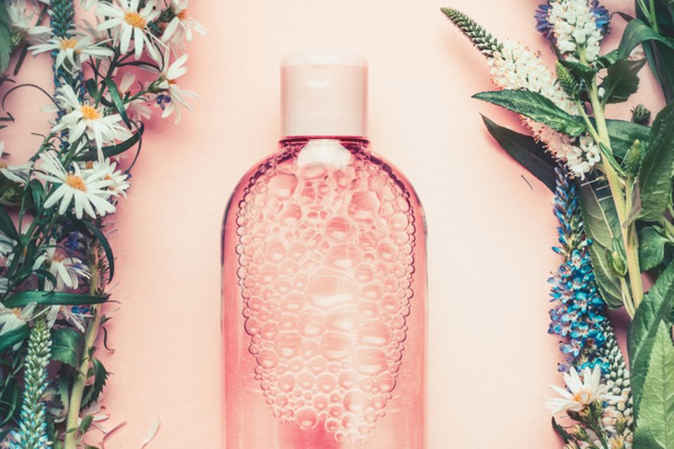 Gesichtswasser selber machen: Flasche mit durchsichtiger Flüssigkeit auf rosa Untergrund, umgeben von Blumen