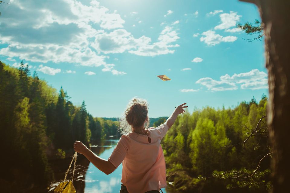 Was ist Glück? Eine junge Frau am Fluss wirft einen Papierflieger in die Luft