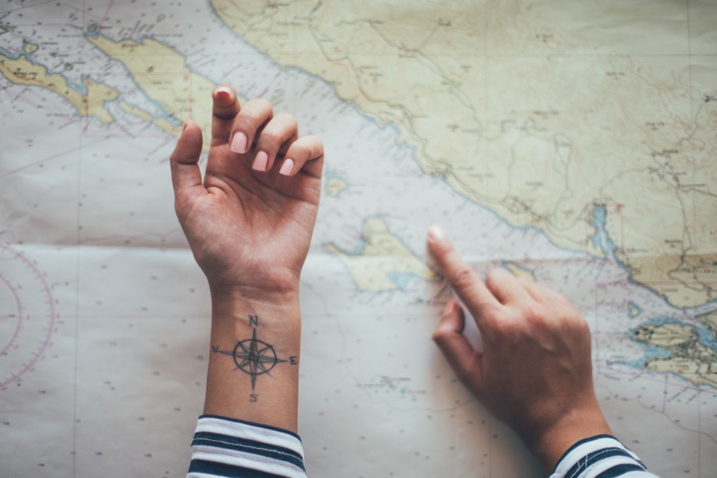Koordinaten-Tattoo: Frauenhand zeigt mit dem Finger auf eine Weltkarte, ihre andere Hand zeigt nach oben