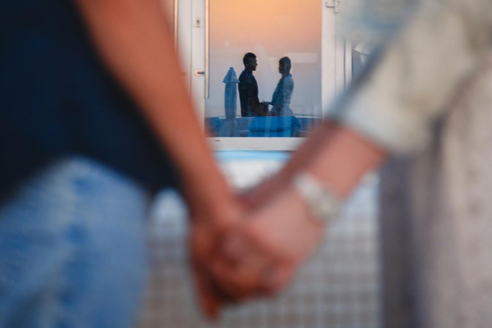 Whisper: Ein Paar steht sich gegenüber, hält sich an den Händen und spiegelt sich in einem  Fenster