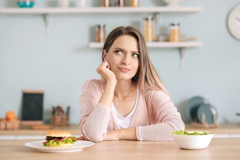 Blähende Lebensmittel: Frau sitzt zwischen Salat und Burger