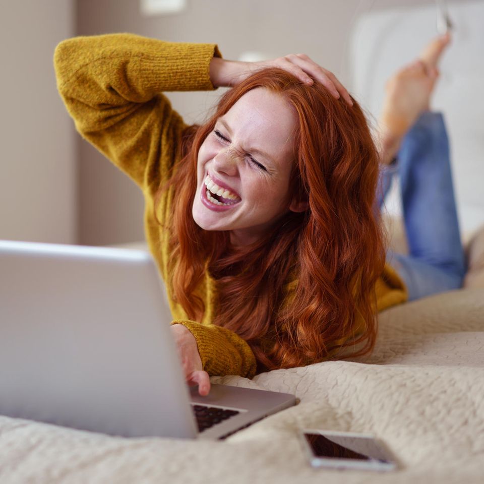 Social Distancing: Eine junge, fröhliche, rothaarige Frau liegt vor ihrem Laptop und lacht