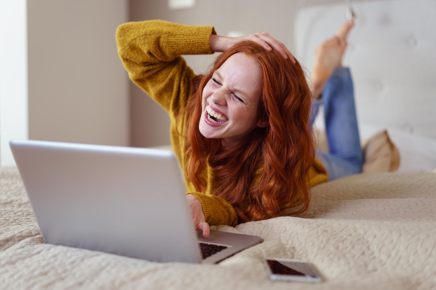 Social Distancing: Eine junge, fröhliche, rothaarige Frau liegt vor ihrem Laptop und lacht