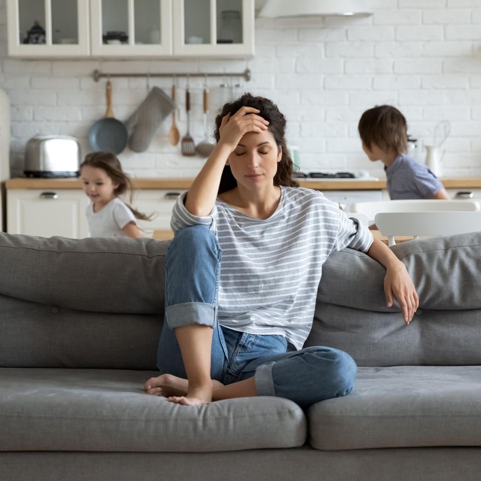 Corona-Lügen: Eine genervte Mutter sitzt auf der Couch, während ihre Kinder hinter ihr toben