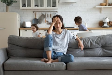 Corona-Lügen: Eine genervte Mutter sitzt auf der Couch, während ihre Kinder hinter ihr toben