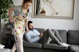 Eine Frau saugt Staub, während ihr Mann auf der Couch sitzt
