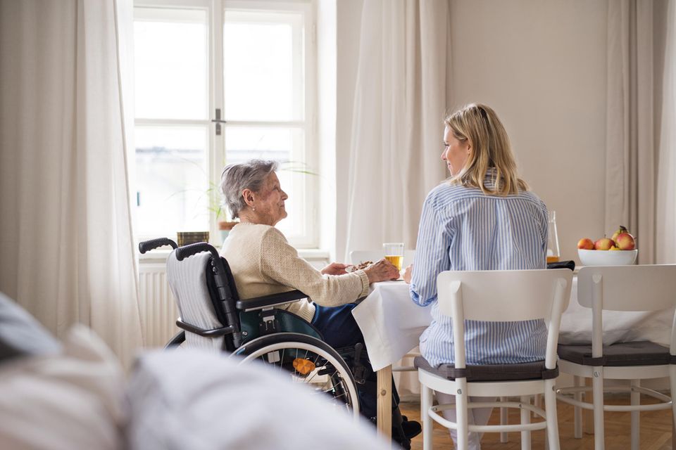 Pflege der Eltern: Ältere Frau im Rollstuhl sitzt mit jüngerer Frau am Tisch