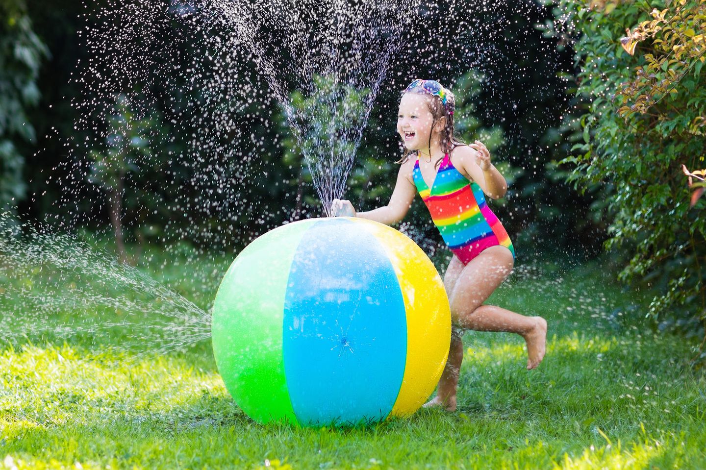 Wasserspielzeug: Mädchen in Badeanzug mit Wasserspielzeug im Garten, bunter Wasserball mit Düsen für Kinder