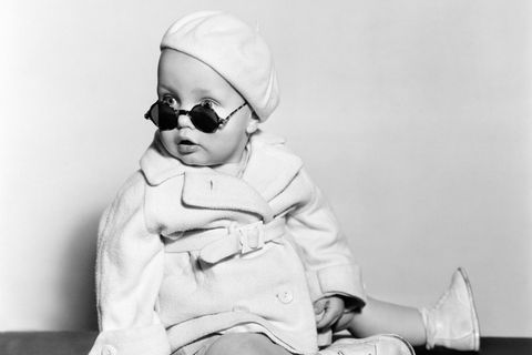 Ein Kleinkind mit einem Mantel bekleidet und einer großen Sonnenbrille sitzt auf dem Boden und schaut in die Kamera