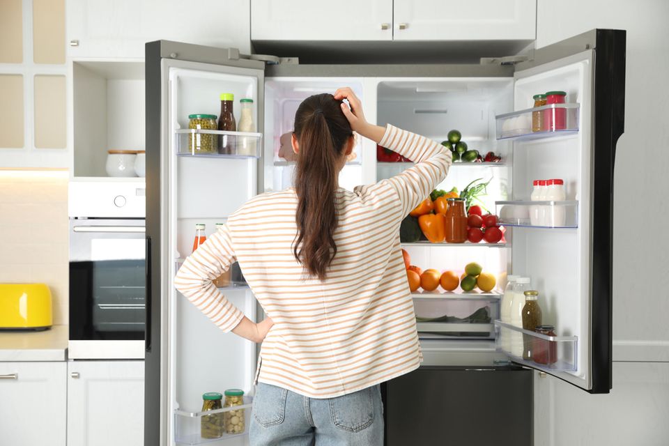 Eine Frau steht vor dem offenen Kühlschrank und kratzt sich am Kopf