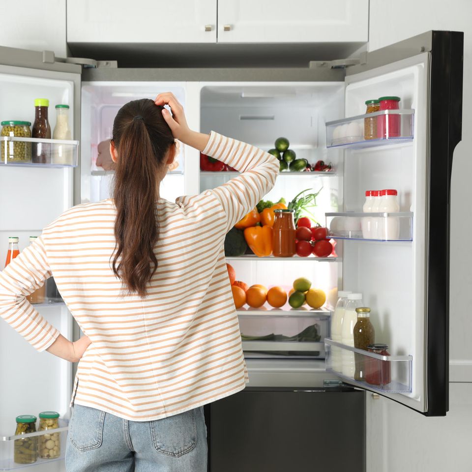 Eine Frau steht vor dem offenen Kühlschrank und kratzt sich am Kopf