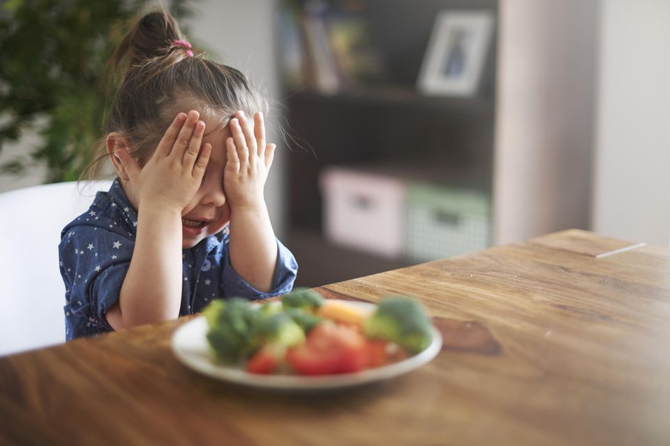 Ein Kind hält sich die Augen zu, während es vor einem Teller Gemüse am Tisch sitzt.