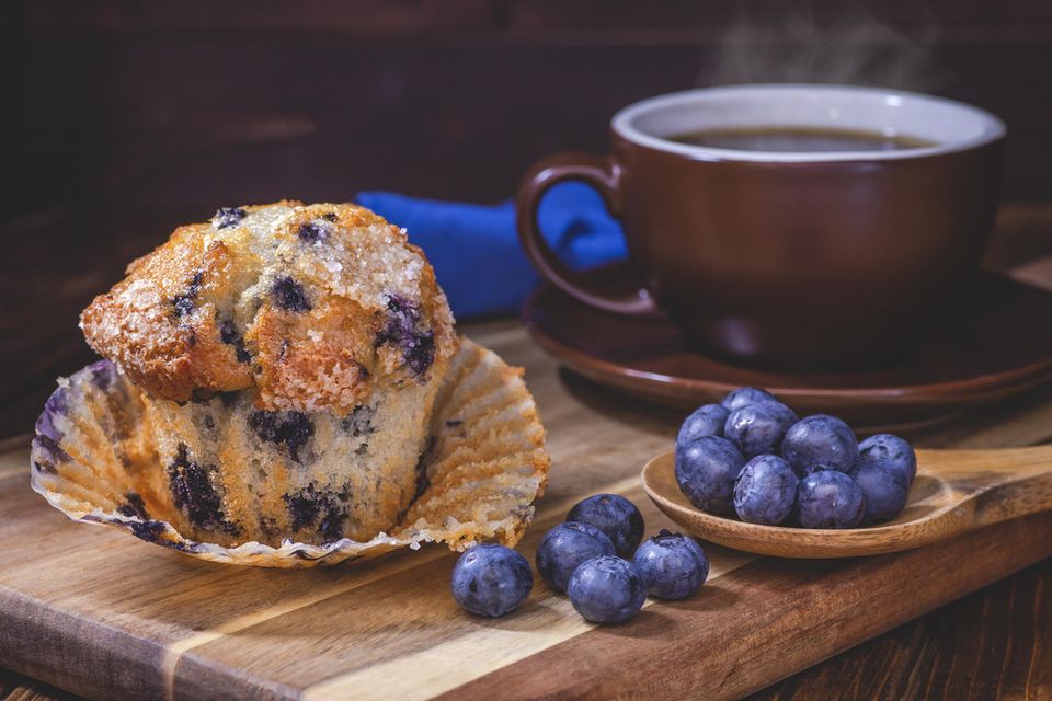 Blaubeer-Muffins mit Blaubeeren in einem Löffel und einer Tasse Kaffee auf einem Holzbrett.