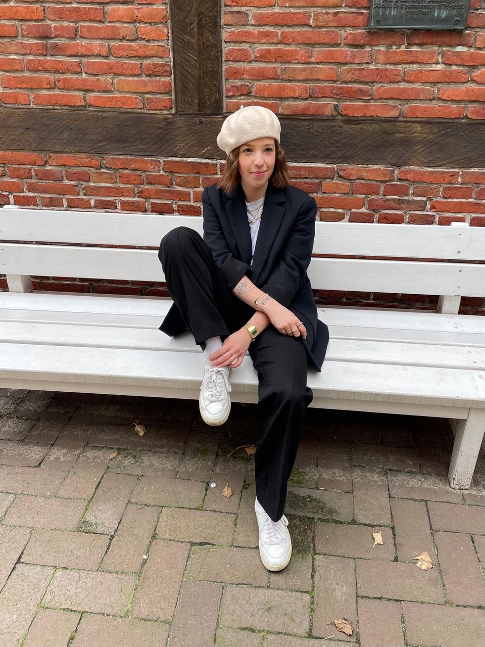 Shopping Queen: Drei Brigitte-Redakteurinnen mit ihrem Style zum Motto "Anzüge" Brünette Frau mit schwarzem Anzug und weißen Turnschuhen und beiger Mütze auf einer weißen Bank