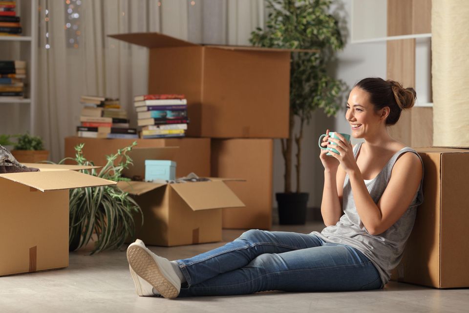 Eine Frau sitzt glücklich allein in ihrer Wohnung und trinkt aus einer Tasse.