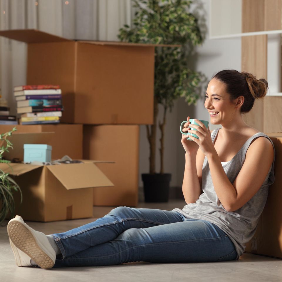 Eine Frau sitzt glücklich allein in ihrer Wohnung und trinkt aus einer Tasse.