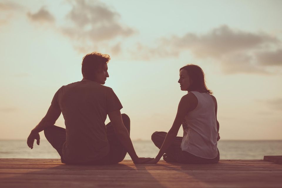 Partner mit Autismus: ein Paar sitzt im Schneidersitz auf einem Steg am Wasser vor einem Sonnenaufgang und sieht sich an.