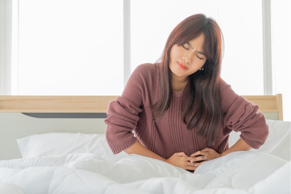 Asiatische Frau mit brünettem Haar in Bett hält sich vor Schmerzen den Bauch