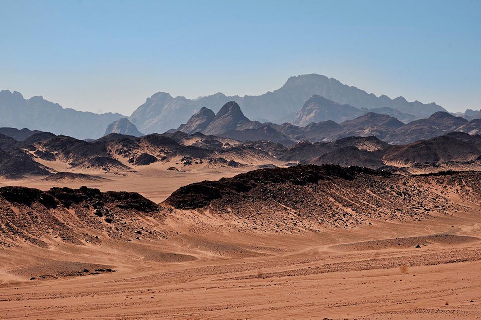 Trans-Reisen: Eine bräunliche Wüstenlandschaft mit zerklüfteten Felsen in der Ferne und blauem Himmel