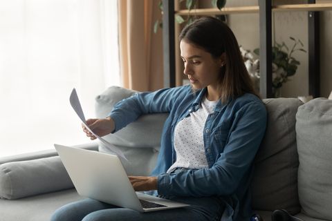 Zusatzversicherungen: Eine junge Frau mit dunklen Haaren sitzt mit ihrem Laptop auf dem Schoß auf einer grauen Couch.