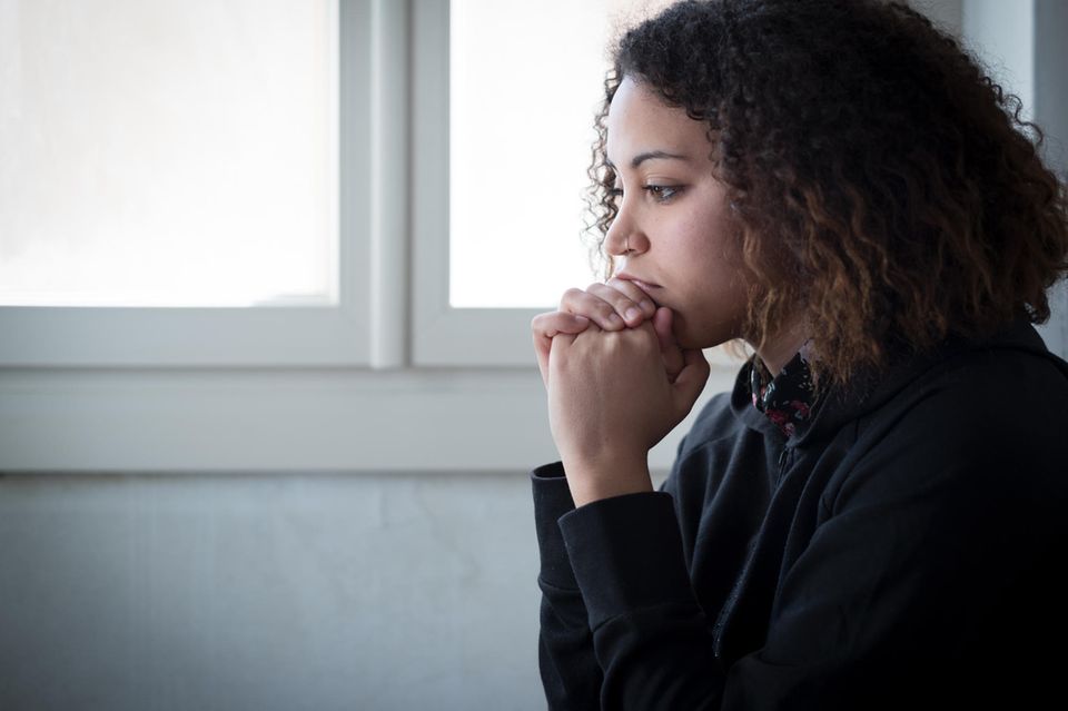Mobbing in der Schule: Eine Teenagerin mit dunklem Pulli und dunklen lockigen Haaren sitzt traurig neben einem Fenster.