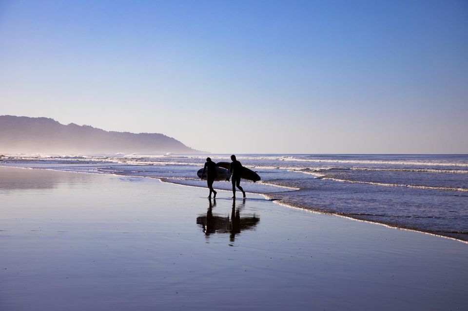 Nicoya: die Silhouetten von zwei Personen mit Surfbrettern, die am Strand gehen.