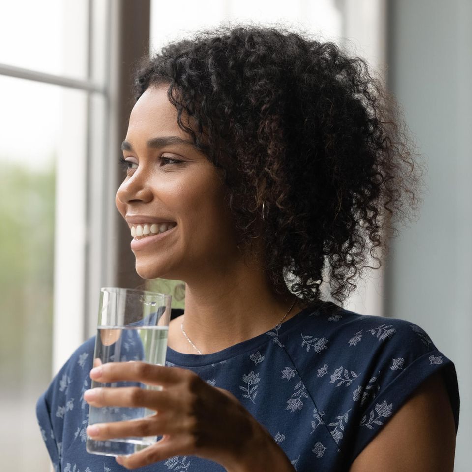Frau trinkt Wasser am Fenster: 5 Gründe, warum du morgens als Erstes ein Glas Wasser trinken solltest