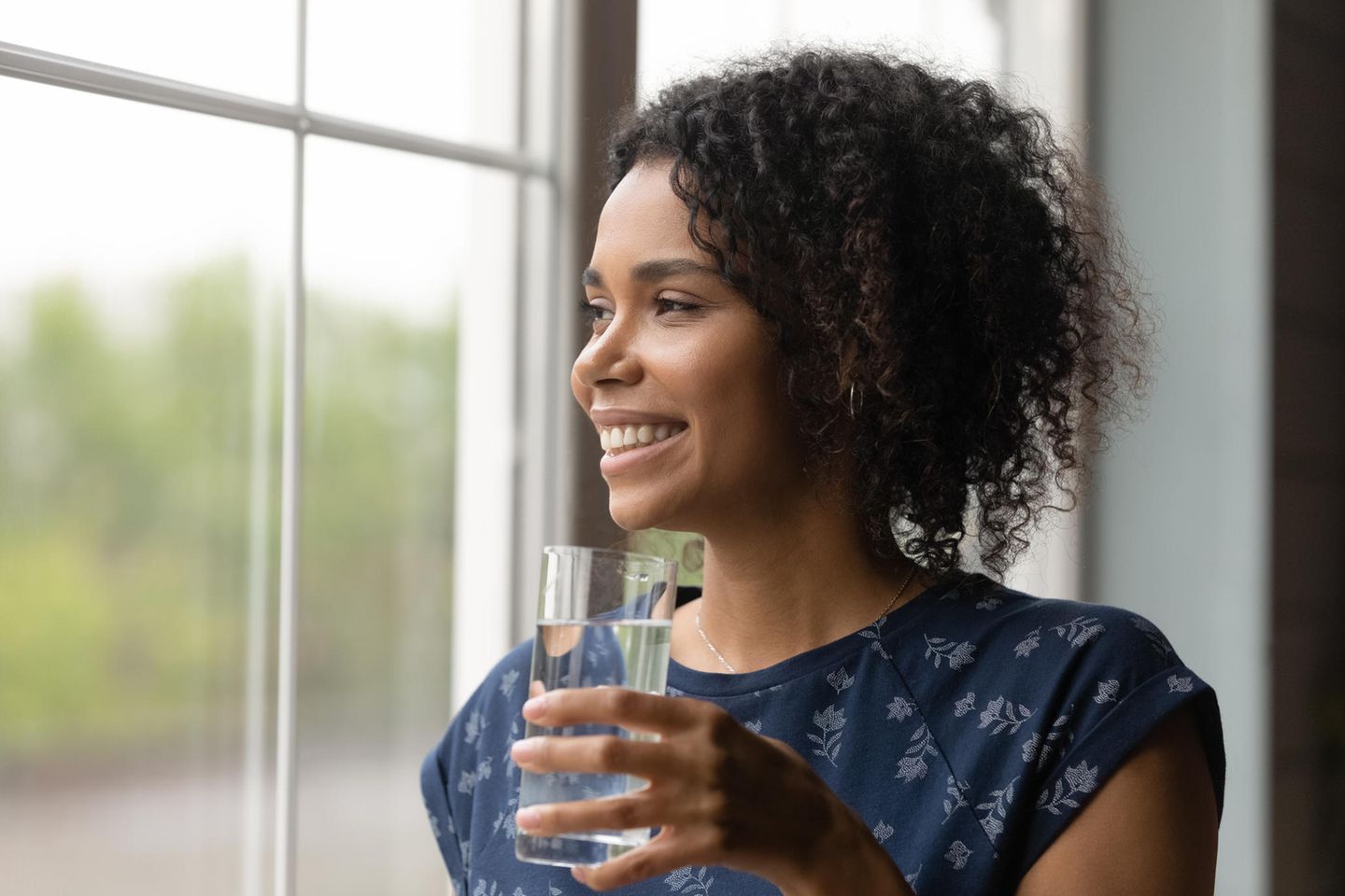 Frau trinkt Wasser am Fenster: 5 Gründe, warum du morgens als Erstes ein Glas Wasser trinken solltest