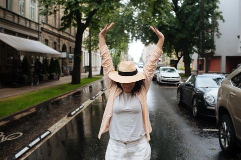 Meike Cornelius: Eine Frau geht auf einer Straße auf die Kamera zu. Sie trägt einen Hut und streckt die Arme in die Luft