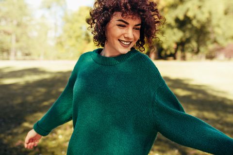 Verenas Selbstversuche: Frau mit braunen Locken und grünem Pullover steht lächelnd auf einer Wiese