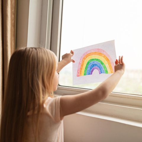 Ein Kind steht am Fenster und hält ein Bild mit einem Regenbogen hoch.