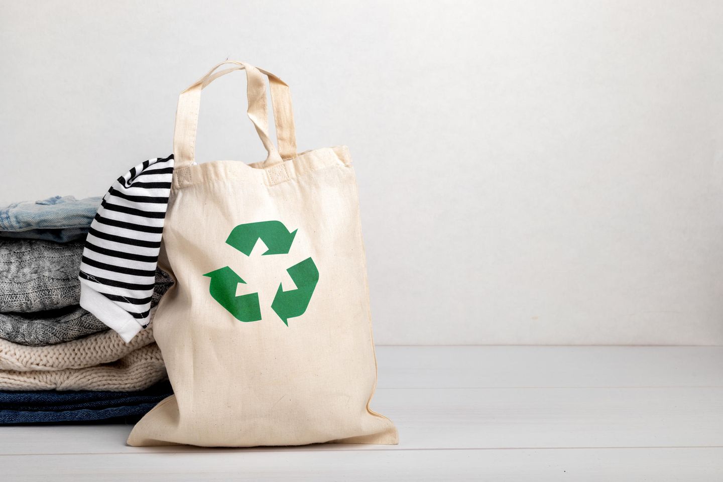 Kleidung recyclen: Ein Stapel Kleidung neben einem Jutebeutel mit einem Recycling Logo