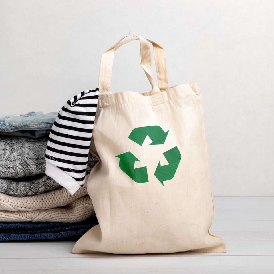 Kleidung recyclen: Ein Stapel Kleidung neben einem Jutebeutel mit einem Recycling Logo